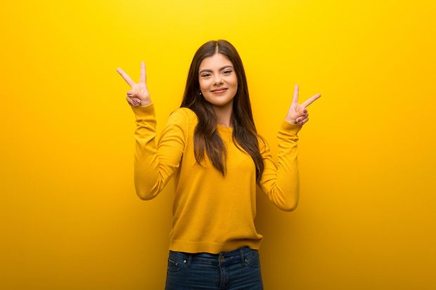 Teenager girl on vibrant yellow background smiling and showing victory sign with both hands